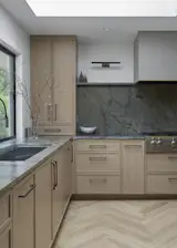 Corner of the remodeled kitchen featuring white oak cabinetry and dramatic stone surfaces that extend into a full-height backsplash. A large 48-inch Kohler sink is centered beneath the window, bringing light across the workspace and anchoring the herringbone wood floor and reconfigured range wall.