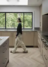 Wide kitchen view showing the 48" sink centered beneath an expanded window that frames the surrounding greenery. The herringbone wood floor and white oak cabinetry create a warm, continuous backdrop as movement through the space highlights the openness of the reconfigured layout.